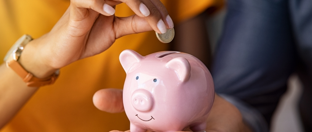 A man holds a small pink piggy bank out as a woman in a yellow shirt puts a coin in.