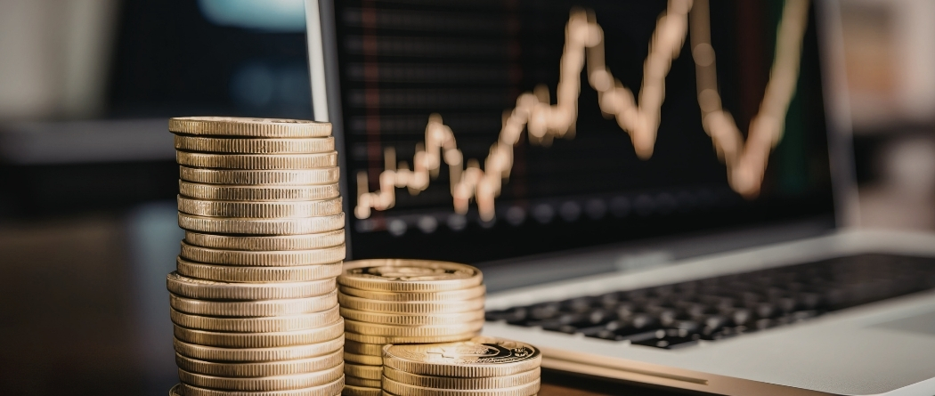 A stack of coins sits in front of a computer with a rising graph on the screen