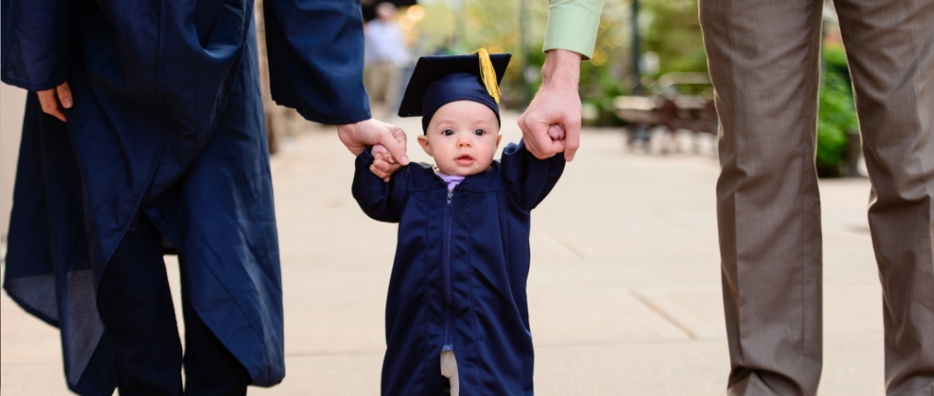 parents holding baby's hand as he walks dressed in a cap and gown.