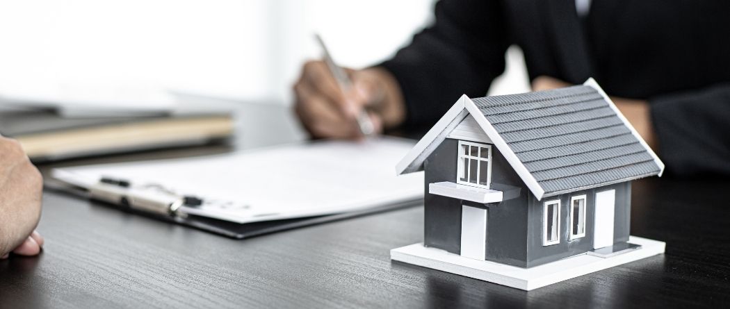 hand completing paperwork with a wooden house sitting on the desk