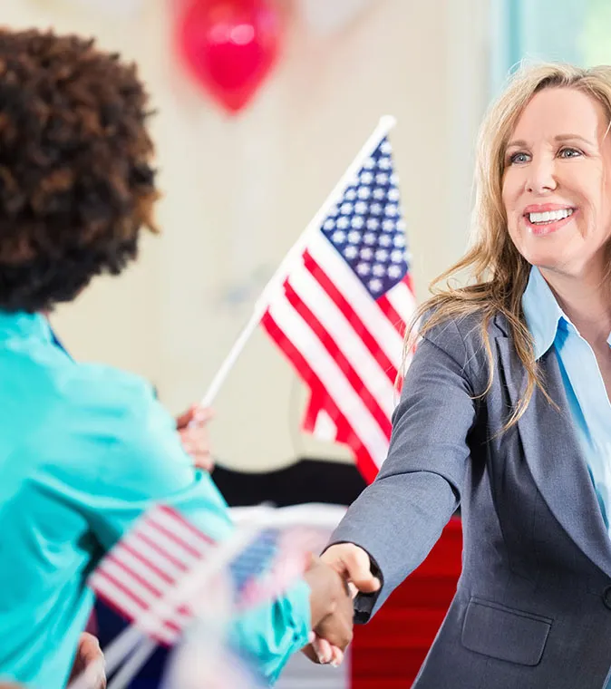 Two women shaking hands with an American flag in the background.