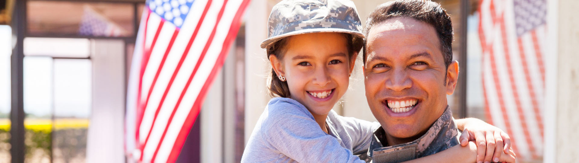Man in military uniform smiling and holding his daughter, who is wearing his helmet.