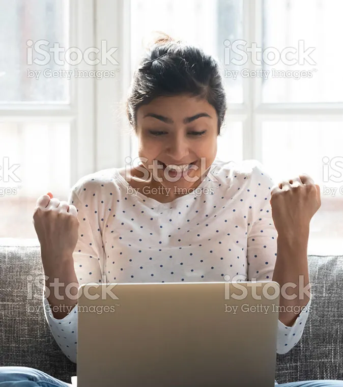 A woman smiling and raising her hands in the air while using a laptop computer.